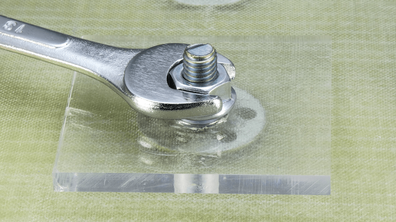 Photograph showing a close-up of a spanner tightening a nut onto a transparent plastic plate. The image depicts the fastening functionality of the nut securing the plate onto a stud-fixing bigHead fastener that is flush embedded within a glass-fibre laminate material.